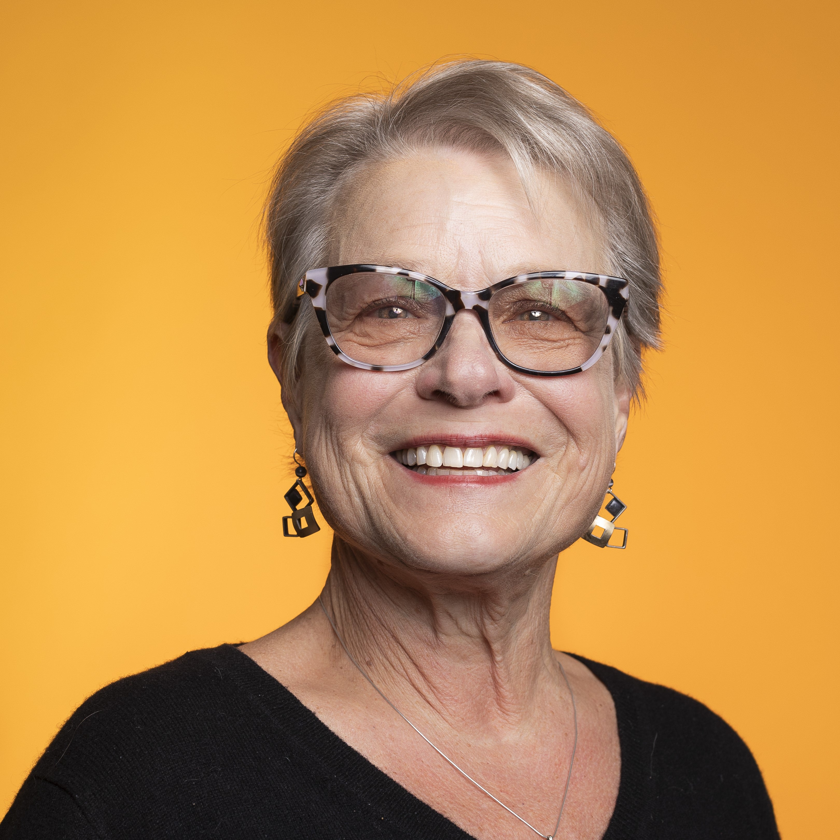 Brenda Major with black top, short blonde hair, and glasses against yellow backdrop
