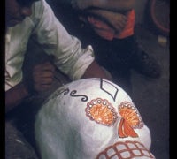 A person hand-painting intricate orange and black decorative designs onto a large white papier-mâché skull.