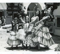 A black and white photograph of four flamenco dancers of different ages posing together in a sunny courtyard. They wear traditional ruffled dresses and flowers in their hair, striking poses with raised arms. The background shows white stucco buildings and