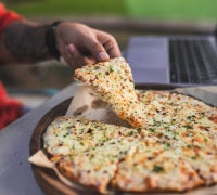 A close-up shot of a person lifting a slice of thin-crust cheese pizza sprinkled with herbs and red pepper flakes from a round wooden platter, with a laptop blurred in the background.