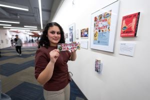 A student poses with a UCSB Reads 2026 bookmark while standing beside a framed promotional poster for UCSB Reads 2026 and a wall-mounted copy of Crying in H Mart that are featured in the Creating Community Through Books exhibition.